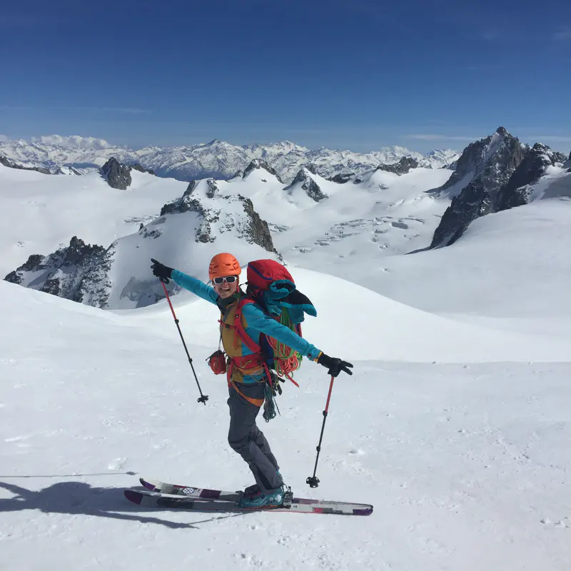 Een skiër in felgekleurde kleding poseert met wijd uitgestrekte armen op een besneeuwde berghelling, omringd door met sneeuw bedekte bergtoppen onder een strakblauwe hemel.